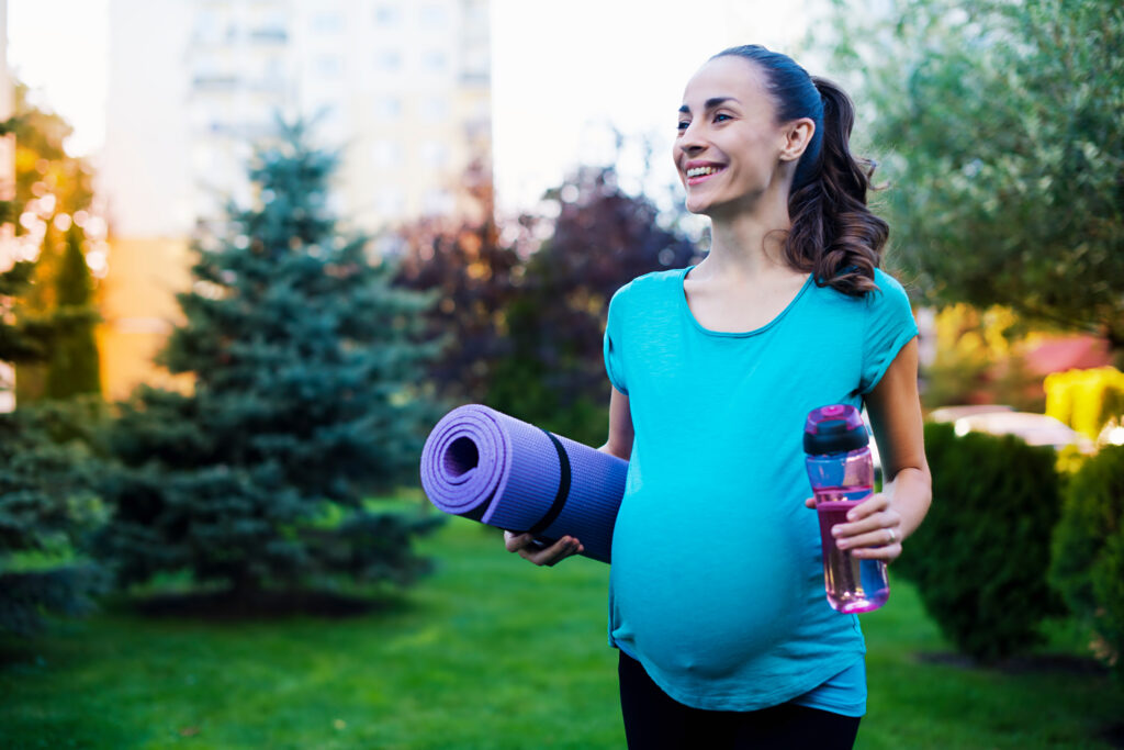 Femme enceinte souriante, tapis de sport à la main, en extérieur